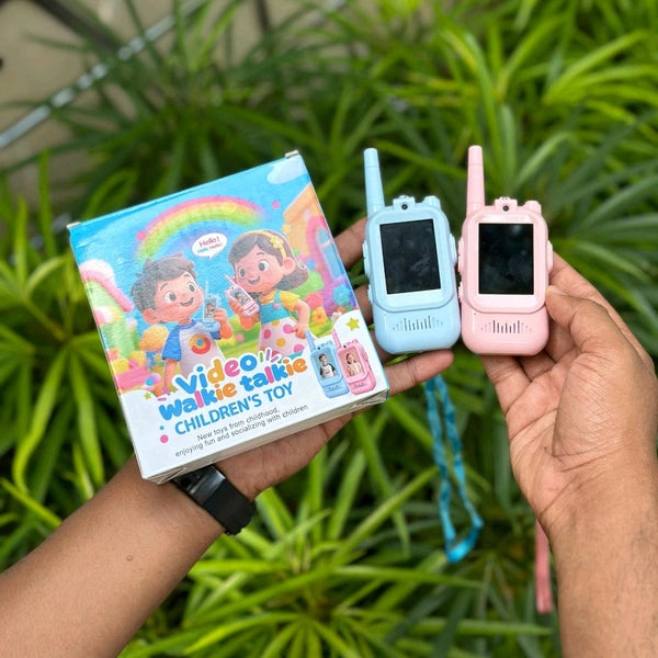 Two toy walkie-talkies, one blue and one pink, held by a person with a colorful box labeled 'Video Walkie Talkie Children's Toy' against a green plant background.